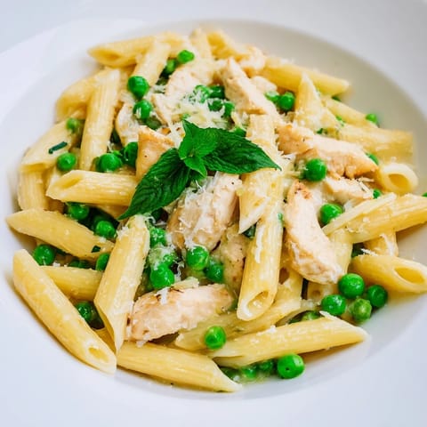 Steaming forks of pea and mint chicken pasta twirl from a bowl, showcasing golden chicken strips, bright green peas, and a glossy Parmesan-lemon sauce that glistens on the noodles.