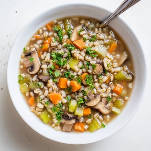 Close-up of homemade mushroom and barley soup, showing golden-brown mushrooms, diced carrots, and fresh herbs floating in a rustic, simmering pot.